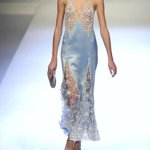 Runway shot of a model wearing a light blue silk slip dress with hand-applied sheer lace bodice panels, featuring a deep V-neckline and a fluid bias-cut silhouette.