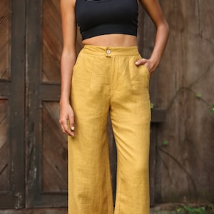 May include: Mustard yellow wide-leg pants with a button closure and pockets. The pants are paired with a black crop top. The pants have a high waist and a relaxed fit. The background is a wooden door.