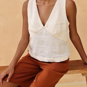 Medium cropped shot of the model seated, focusing on the distinct paneled seam construction underneath the deep V-neckline of the cream linen tank top.