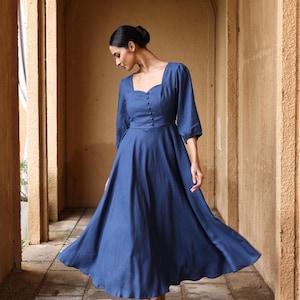 Full-length front three-quarter view of a woman walking gracefully in an outdoor stone corridor, wearing a slate blue linen maxi dress with a sweetheart neckline, a buttoned bodice, and three-quarter puff sleeves.