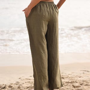 Side-profile full-body view of the model looking out at the ocean, showing how the loose, flowing linen fabric drapes elegantly in natural daylight on the beach.