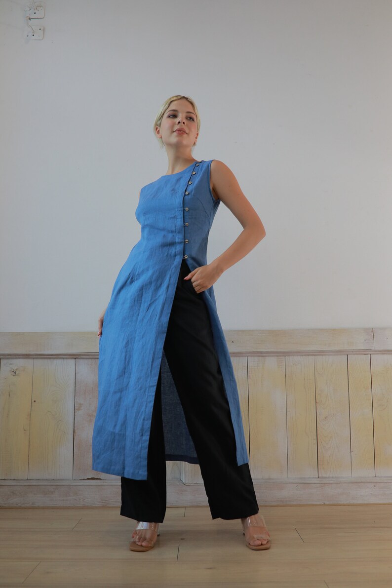Full-length side-profile shot of a blonde model posing with one hand on her hip, wearing a sleeveless denim-blue linen tunic top with a high front slit, paired with wide-leg black trousers.