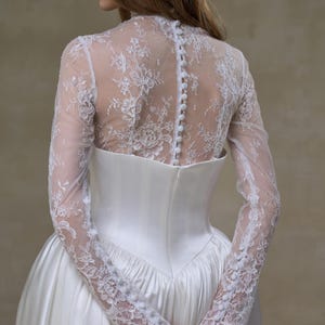 May include: A white gown featuring a high-neck lace bodice with long sleeves and a row of small fabric-covered buttons running down the center back. The bodice transitions into a smooth, solid white satin skirt with a structured waistline.
