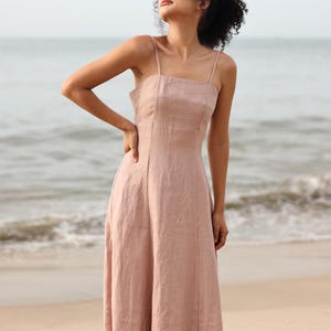 Waist-up shot of a woman looking up and off-camera, wearing a dusty pink linen slip dress with thin straps, standing against a backdrop of a calm ocean.