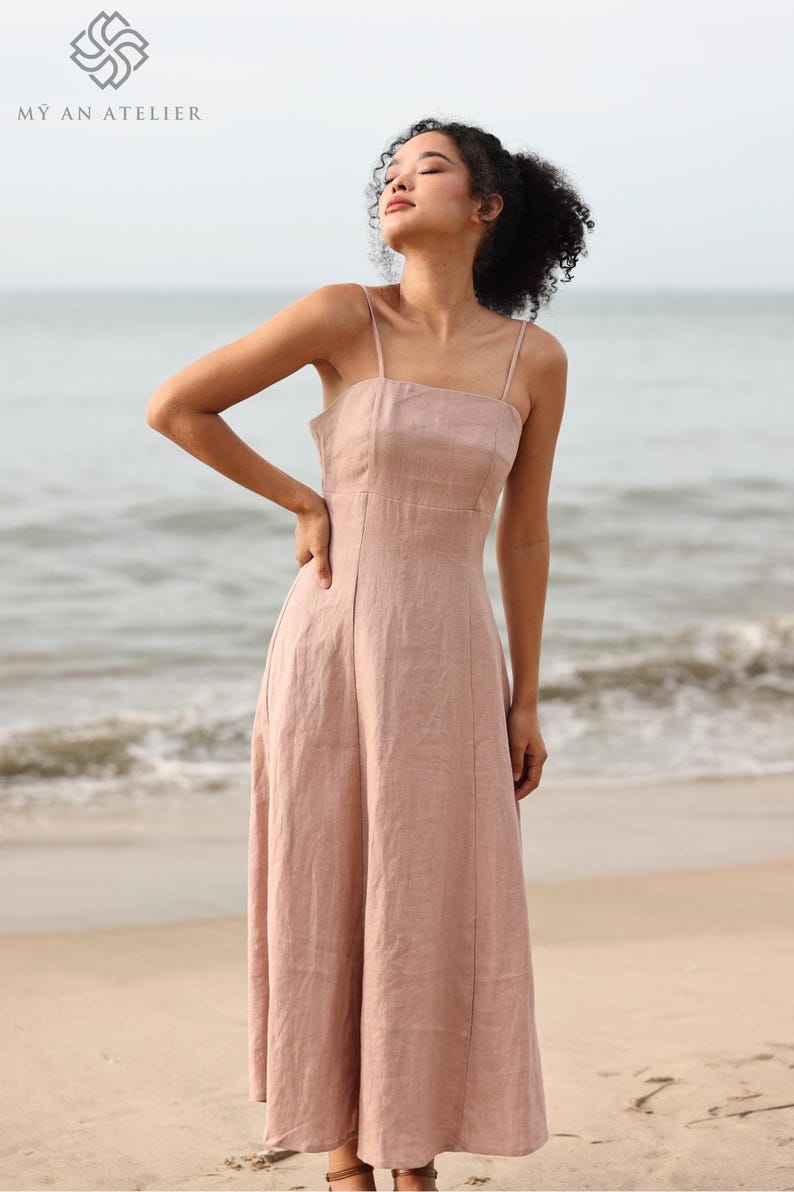 Waist-up shot of a woman looking up and off-camera, wearing a dusty pink linen slip dress with thin straps, standing against a backdrop of a calm ocean.
