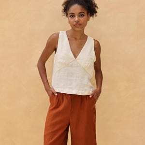 Full-length front shot of the model standing with both hands tucked into the pockets of her terracotta brown wide-leg trousers, displaying a complete minimalist summer lifestyle outfit.