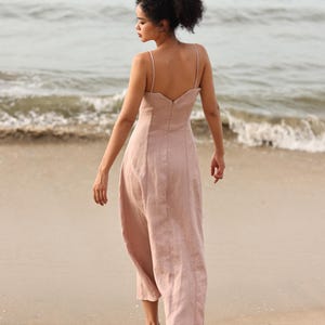 Full-length back view of a woman walking away on a sandy beach toward the ocean waves, showcasing the low-cut back design of a dusty pink linen maxi dress.
