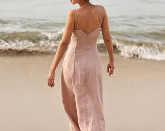 May include: A light pink dress with thin straps and a V-neck is worn on a beach. The dress has a fitted bodice and a flowing skirt. The person is wearing brown sandals. The logo "MY AN ATELIER" is in the top left corner.