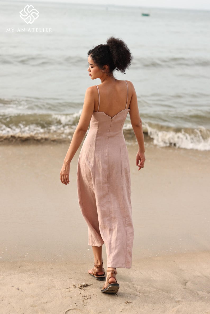 Full-length back view of a woman walking away on a sandy beach toward the ocean waves, showcasing the low-cut back design of a dusty pink linen maxi dress.