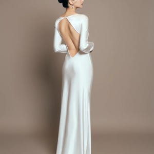 May include: A long, white satin gown with long sleeves and a dramatic open back. The dress features a high neckline in the front and a flowing skirt that trails behind. The model has dark hair styled in a bun.