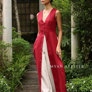 Puede incluir: Una túnica roja sin mangas, abotonada, con cuello en V, combinada con pantalones de pierna ancha color crema. El conjunto es de MYAN ATELIER. La modelo está de pie en una pasarela de madera rodeada de vegetación.