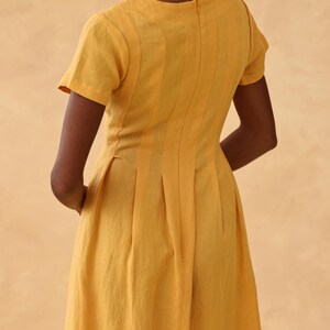 Medium back and side three-quarter view of a woman, highlighting the structured vertical seams, darts, and inverted pleats on the back waistline of a yellow linen dress.