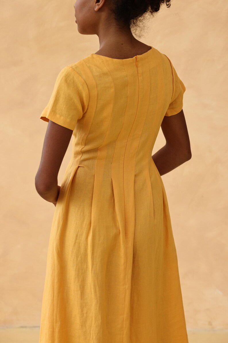 Medium back and side three-quarter view of a woman, highlighting the structured vertical seams, darts, and inverted pleats on the back waistline of a yellow linen dress.