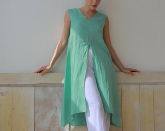 May include: A woman wearing a light green sleeveless tunic with a front slit and white pants. She is standing in front of a light wood panel wall.