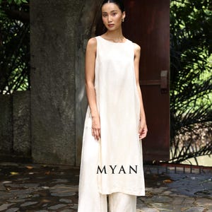 Könnte beinhalten: Eine Frau trägt ein cremefarbenes ärmelloses Oberteil und eine Hose mit weitem Bein. Das Outfit besteht aus einem leichten, fließenden Stoff. Das Wort "MYAN" ist auf das Oberteil gedruckt. Der Hintergrund umfasst eine Steinmauer, eine Holztür und Grün.