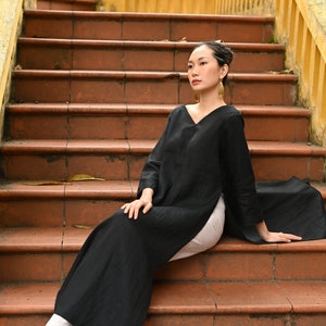 The model is elegantly seated on wide terracotta outdoor stairs, showcasing the side profile and long flowy drape of the black tunic and white trousers combination.
