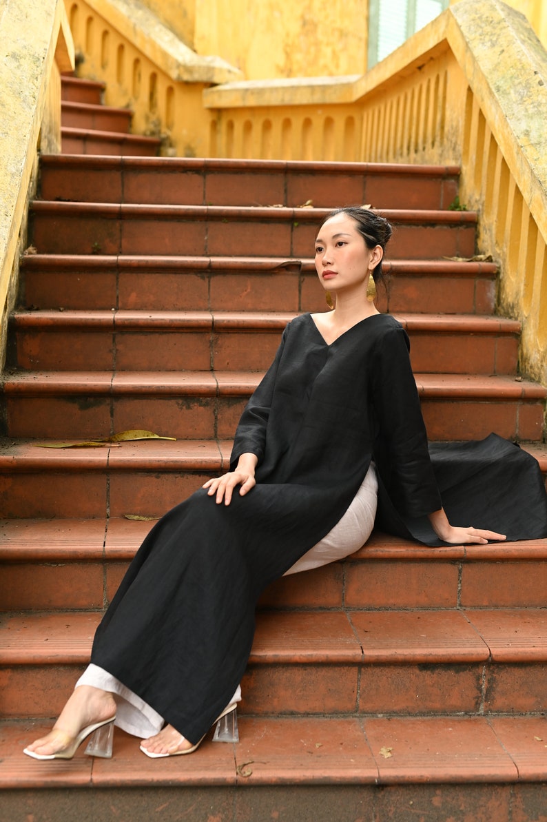 The model is elegantly seated on wide terracotta outdoor stairs, showcasing the side profile and long flowy drape of the black tunic and white trousers combination.