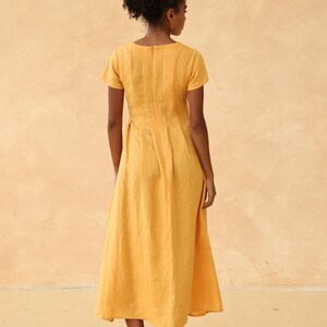 Full-length back view of a woman standing straight, showcasing the elegant back tailoring, center zipper closure, and flowing silhouette of a mustard yellow linen maxi dress.