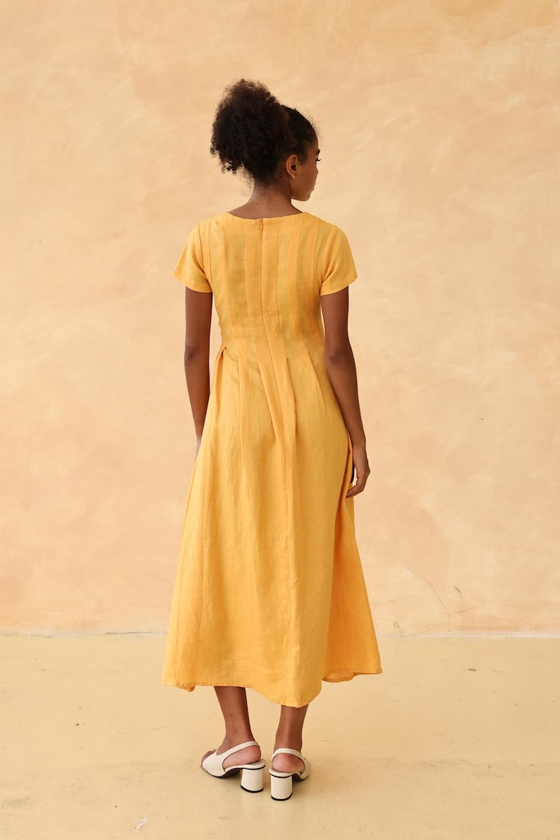 Full-length back view of a woman standing straight, showcasing the elegant back tailoring, center zipper closure, and flowing silhouette of a mustard yellow linen maxi dress.