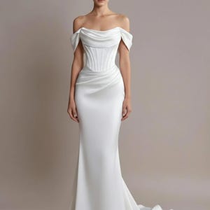 Op de afbeelding: Een witte trouwjurk met blote schouders, een nauwsluitend lijfje en een zwierige rok. De jurk heeft een top in korsetstijl en een sleep. De stof lijkt een glad, satijnachtig materiaal te zijn, wat een elegante silhouet creëert.