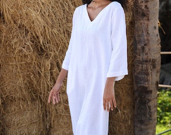 May include: A white linen jumpsuit with a V-neck and three-quarter sleeves. The jumpsuit has a relaxed fit and is suitable for warm weather. The model is standing in front of a hay bale and a palm tree.