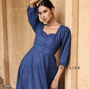Close-up front view from the waist up of a woman posing with one hand near her head against a stone wall, focusing on the romantic sweetheart neckline and elegant three-quarter puff sleeves of a blue linen dress.