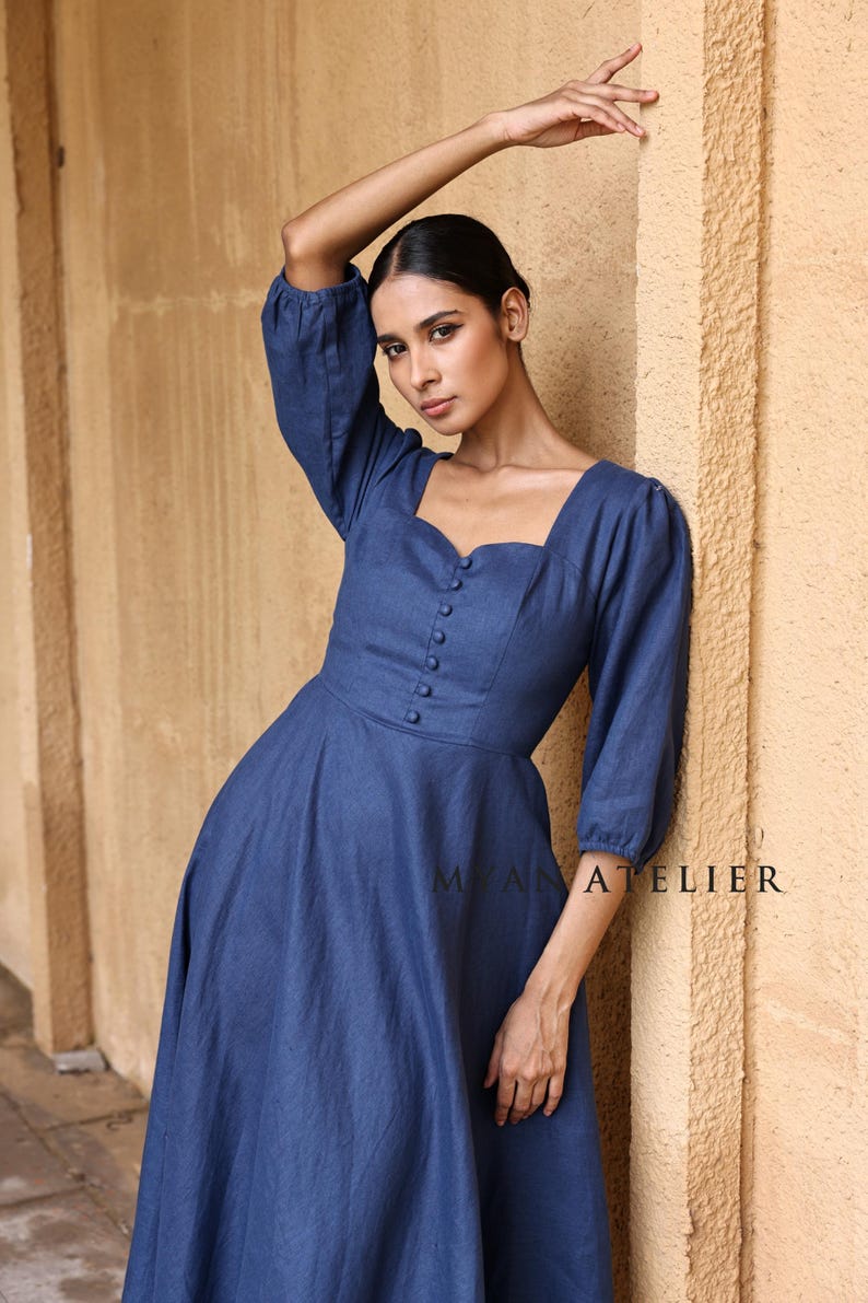 Close-up front view from the waist up of a woman posing with one hand near her head against a stone wall, focusing on the romantic sweetheart neckline and elegant three-quarter puff sleeves of a blue linen dress.