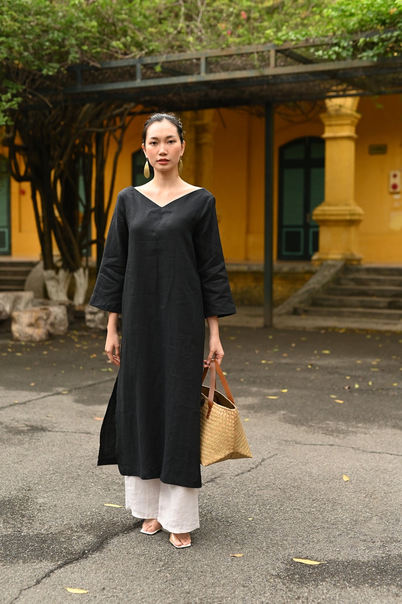 Full-length front shot of the model standing on a paved outdoor pathway, wearing the black V-neck linen tunic and white wide-leg pants.