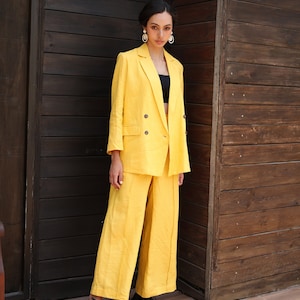 May include: A woman wearing a bright yellow linen suit, consisting of a double-breasted blazer and wide-leg pants. The suit is paired with a black top and black shoes. The background is a dark wooden wall.