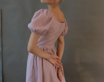 May include: A light pink dress with short, puffed sleeves and a fitted bodice. The dress has a flowing skirt and a scoop neckline. The model is wearing the dress and has her hands crossed behind her back. The background is a solid gray.
