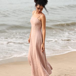 Full-length front view of a woman standing on the beach near the water's edge, wearing a long, flowing dusty pink linen maxi dress with thin straps.