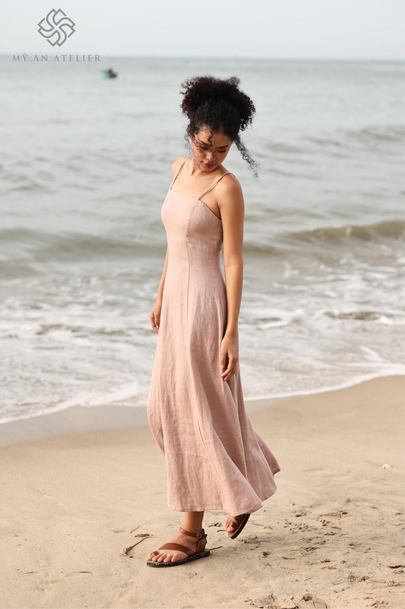 Full-length front view of a woman standing on the beach near the water's edge, wearing a long, flowing dusty pink linen maxi dress with thin straps.