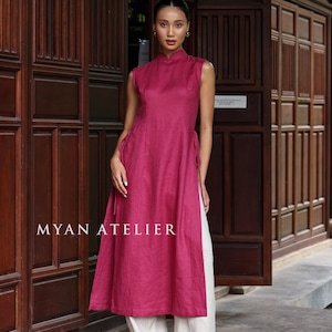 May include: A sleeveless, magenta-colored tunic dress with a mandarin collar and side slits, paired with cream-colored linen pants. The dress features tie details at the waist. The text "MYAN ATELIER" is visible on the front of the dress. The outfit is suitable for warm weather.