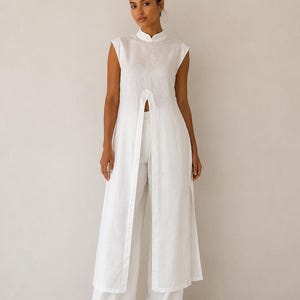 May include: A white linen outfit featuring a sleeveless tunic with a mandarin collar and a front slit, paired with wide-leg pants. The outfit is made of a lightweight fabric, perfect for warm weather. The model is wearing the outfit.