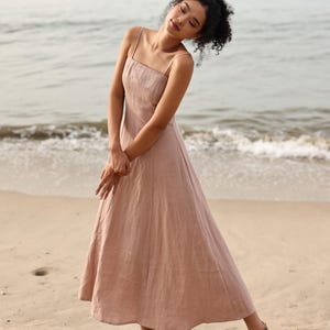 Full-length shot of a woman tilting her head and posing in a dusty pink or light mauve linen maxi dress with thin shoulder straps on a sandy beach with ocean waves in the background.