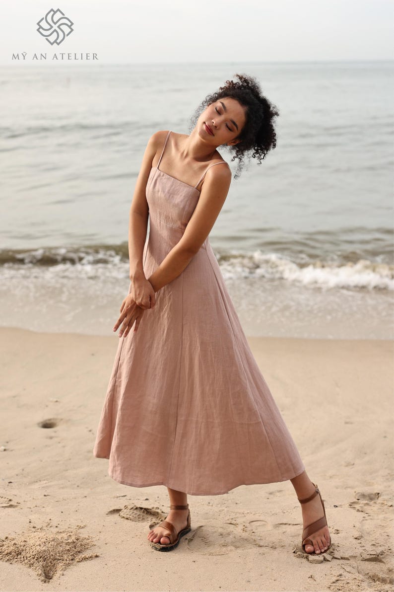 Full-length shot of a woman tilting her head and posing in a dusty pink or light mauve linen maxi dress with thin shoulder straps on a sandy beach with ocean waves in the background.