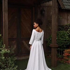 A full-length rear view of the model standing before a rustic wooden cabin. The shot captures the elegant silhouette from behind, featuring a modest mid-high back neckline, a neatly fitted waist, and a flowing A-line skirt that extends into a graceful, subtle floor-sweeping train.