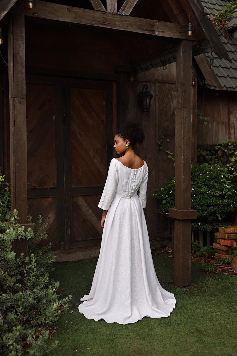 A full-length rear view of the model standing before a rustic wooden cabin. The shot captures the elegant silhouette from behind, featuring a modest mid-high back neckline, a neatly fitted waist, and a flowing A-line skirt that extends into a graceful, subtle floor-sweeping train.