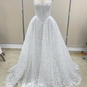 May include: A floor-length white lace gown featuring a strapless sweetheart neckline, a fitted bodice with a vertical row of small fabric-covered buttons down the center, and a full, voluminous skirt with a scalloped lace hemline on a mannequin.