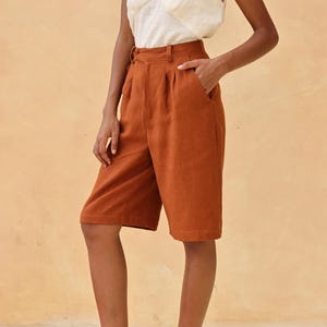 以下が含まれることがあります： 女性は、白いノースリーブのトップスとさび色のリネンショーツを着用しています。彼女は茶色の革のサンダルを履いています。