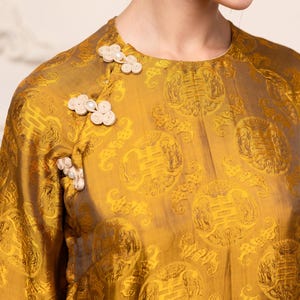 May include: Mustard yellow silk blouse with a repeating pattern and decorative floral buttons. The blouse has long sleeves and a round neckline. The text "MYAN ATELIER" is visible at the bottom of the image.