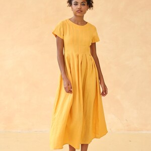 Full-length front shot of a woman looking forward with a relaxed posture, modeling a mustard yellow linen maxi dress with short sleeves against a warm solid backdrop.