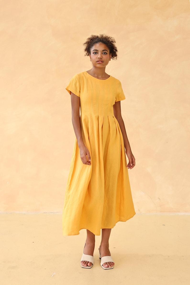 Full-length front shot of a woman looking forward with a relaxed posture, modeling a mustard yellow linen maxi dress with short sleeves against a warm solid backdrop.