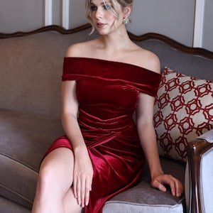 Puede incluir: Vestido de terciopelo rojo oscuro con hombros descubiertos y abertura alta. El vestido tiene la cintura fruncida y cae con elegancia. La modelo está sentada en un sofá de terciopelo gris. Es una prenda formal.