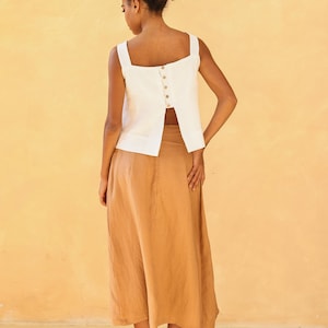 A final full-length rear view of the model standing with her back to the camera. This shot highlights the elegant movement of the split-back hemline opening over the tan linen skirt, anchored by the row of natural buttons running down the center spine.