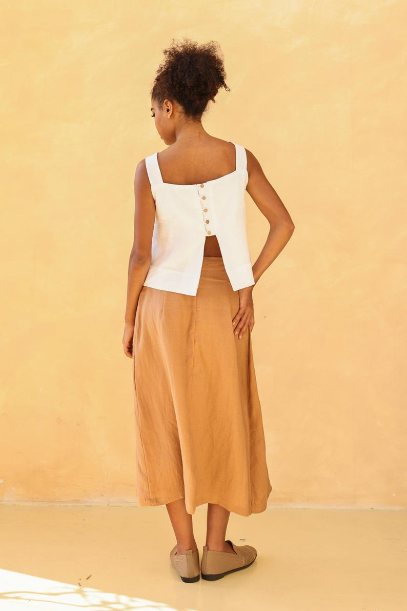 A final full-length rear view of the model standing with her back to the camera. This shot highlights the elegant movement of the split-back hemline opening over the tan linen skirt, anchored by the row of natural buttons running down the center spine.