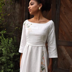 A medium shot of the model looking to the side, highlighting the romantic and artisanal aesthetic of the dress. The image displays the beautifully framed collarbones via the wide boat neckline, the artisanal shoulder embroidery, and the clean French seam construction along the fitted bodice.