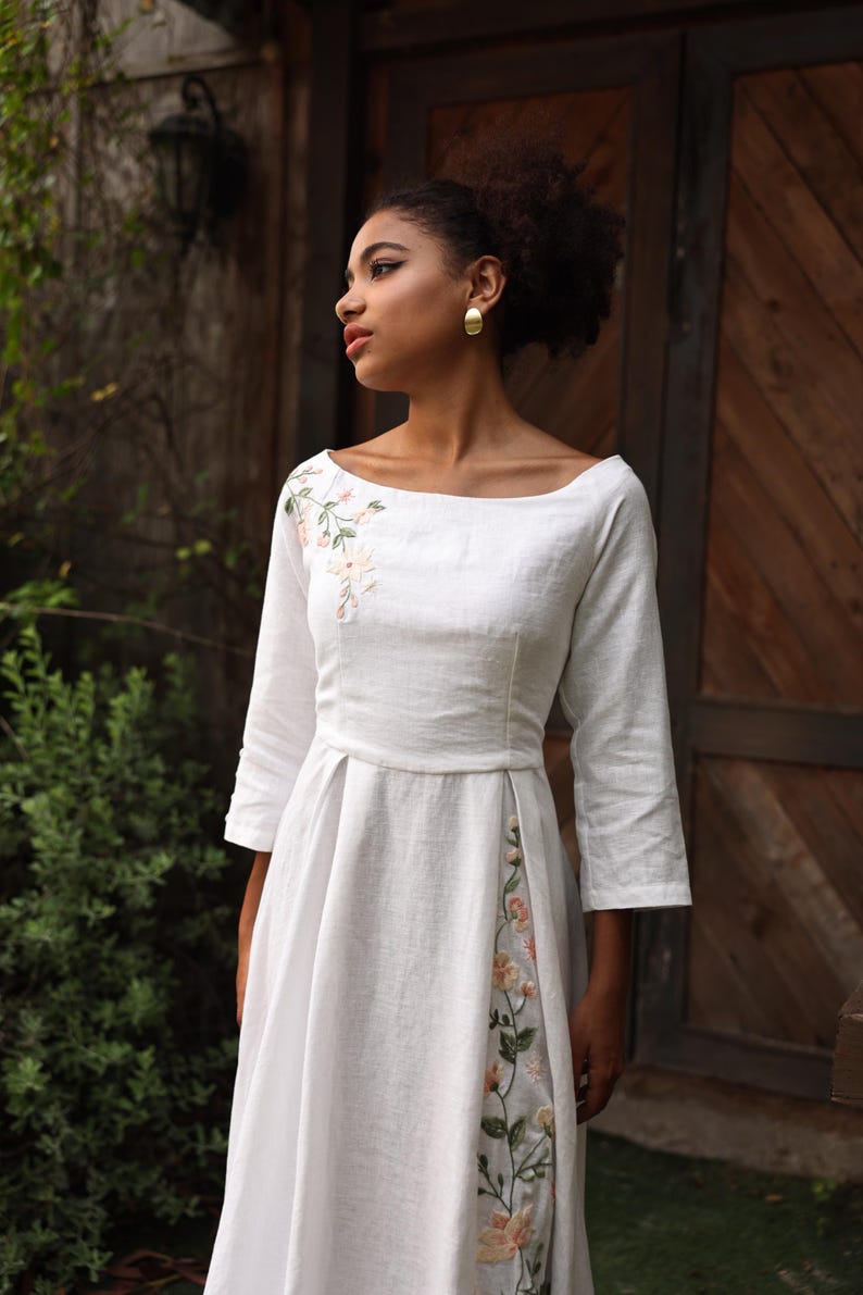 A medium shot of the model looking to the side, highlighting the romantic and artisanal aesthetic of the dress. The image displays the beautifully framed collarbones via the wide boat neckline, the artisanal shoulder embroidery, and the clean French seam construction along the fitted bodice.