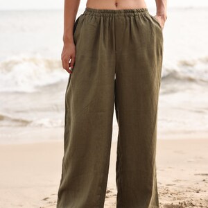 Full-length front shot of the model standing on a sandy beach shore, showcasing the same wide-leg trousers design in an olive green colorway, paired with a black crop top for a beach resort style.