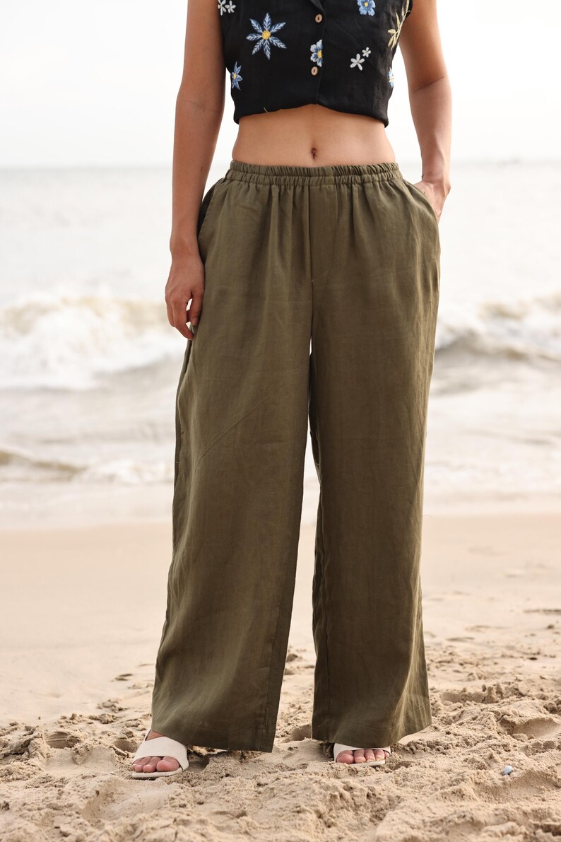 Full-length front shot of the model standing on a sandy beach shore, showcasing the same wide-leg trousers design in an olive green colorway, paired with a black crop top for a beach resort style.